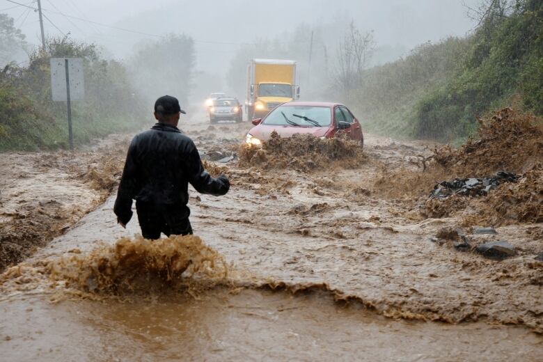 A man in a baseball cap with his back to the camera stands in muddy waters up his legs as motorists approach in a storm.