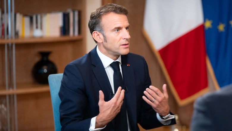 Frances President Emmanuel Macron sits in a chair wearing a blue suit as he is interviewed by CBCs Chief Political Correspondent Rosemary Barton and Radio-Canada's Parliamentary Bureau Chief Louis Blouin who are off camera.