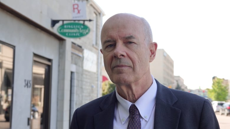 An older man wearing a suit stands in front of a grey brick law office.