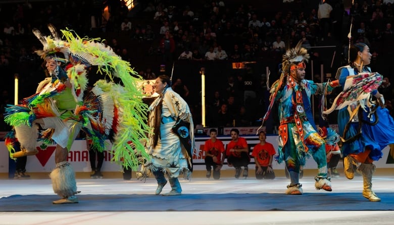 Four people in colourful Indigenous regalia dance on a mat on the ice of a darkened hockey stadium.