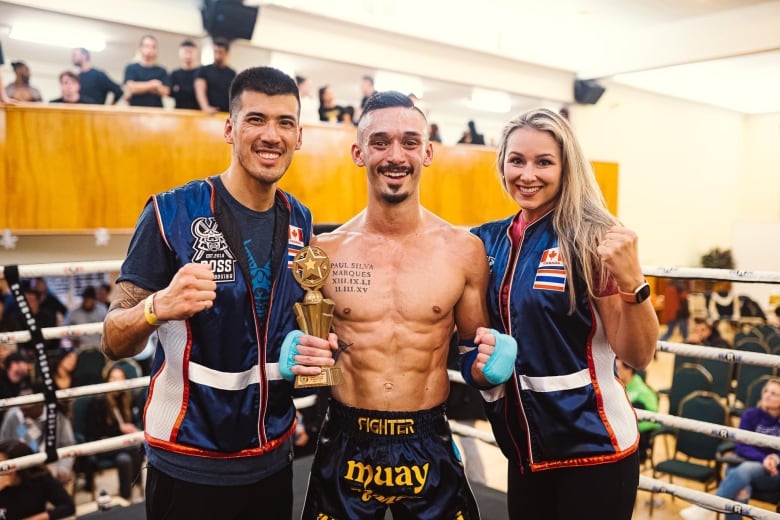 Two men and a woman stand in a boxing ring smiling.