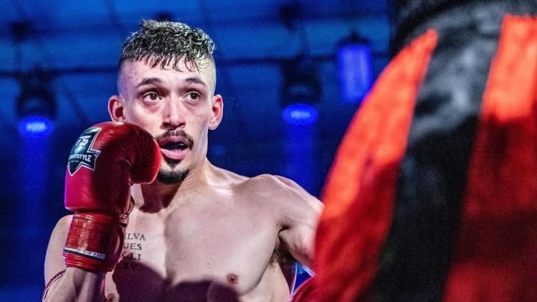 A man with light skin and dark hair stands with a gloved fist in a boxing ring.