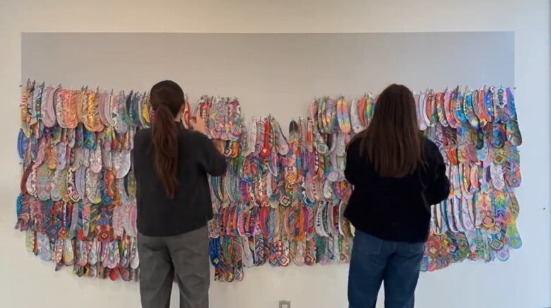 Rows of colourful coloured paper feathers hang on a white wall. Too people are seen from behind hanging more of them.