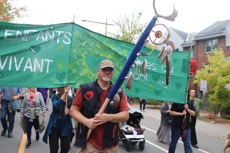 A man in a black vest and orange t-shirt participates in a march. He holds a staff decorated with antlers and dream catchers. A group of people carrying a large green banner walk behind him. 