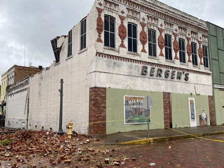 A brick building that has its upper story wall torn down, there are mounds of bricks on the street below.