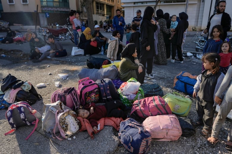 A child sleeps on the ground amid a pile of backpacks as others sit and mingle.