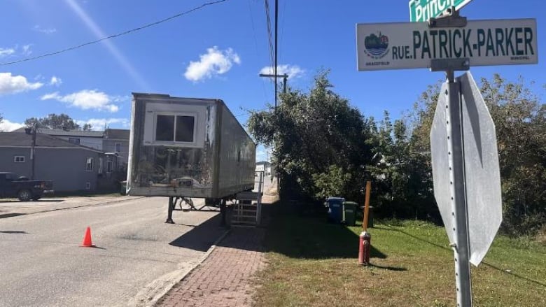 A street on a summer day with a trailer on the side of the road and an orange traffic cone near it.