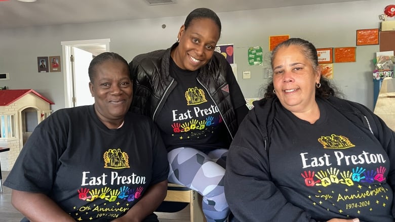 Three women pose for a photo. They are all wearing black t-shirts that say 