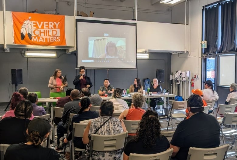 People sitting looking toward a woman holding a mic. An orange flag is hanging over the rafters of the indoor building, with the words 'Every Child Matters'