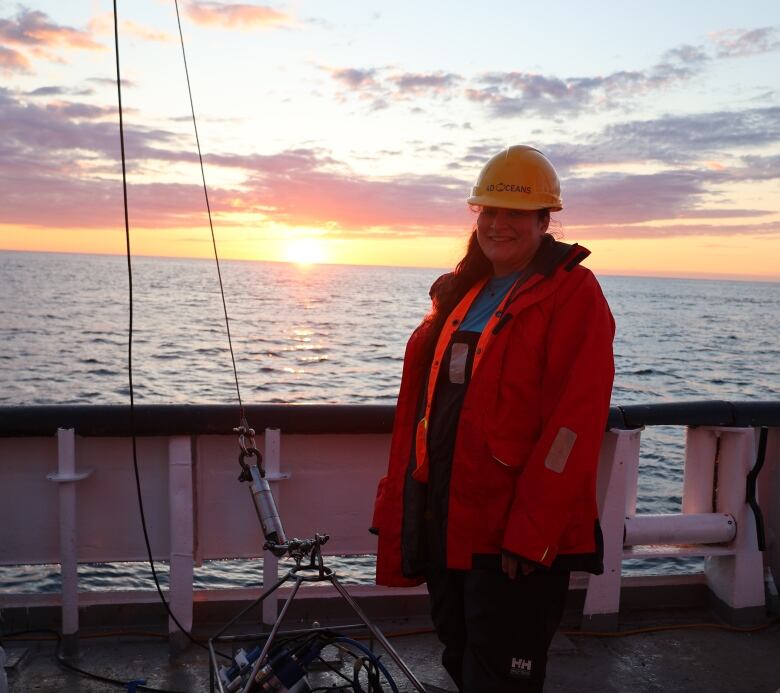 A figure standing on the deck of a boat wearing a large coat. Next to her is a camera in a box.