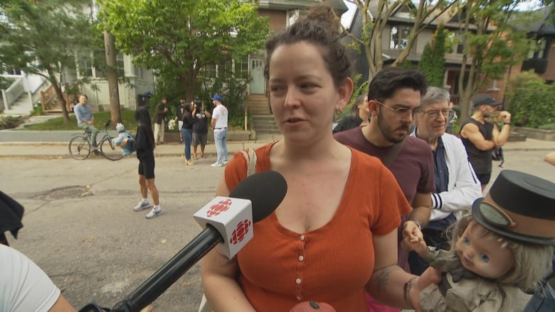 A woman, visible from the chest up, speaks into a reporter's microphone on a residential city street on a sunny afternoon. She is holding a doll in a top hat.