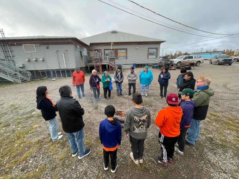 People from kids to elders stand in a circle around a fire, many of them wearing orange shirts