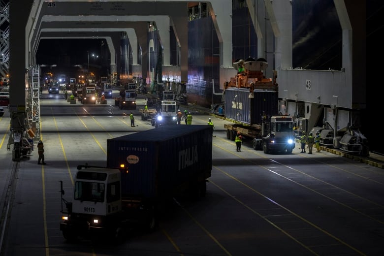 A crane places a container on the back of a truck as other vehicles navigate a dock.