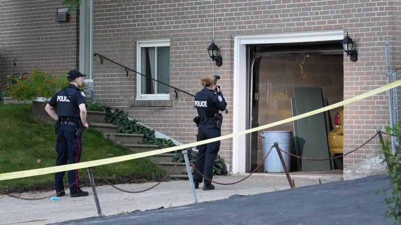 Two police officers, one male, one female, stand in a residential driveway that's taped off from the street. The female officer is photographing an open garage. It is late evening and sunny.