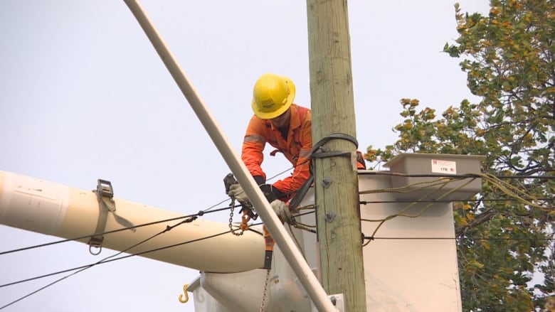 A man in a yellow hardhat and orange jumpsuit stands in the bucket of a crane and works on wires.