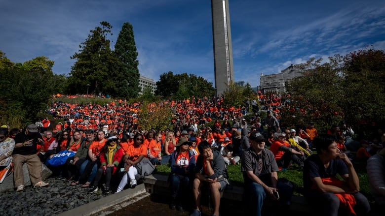 A sea of people wearing orange shirts sit on a lawn during a sunny day.