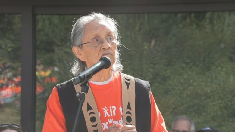 An older First Nations man wearing orange looks up while speaking at a mic.