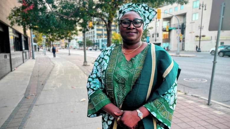 A woman in a green traditional Nigerian outfit stands on a sidewalk and smiles