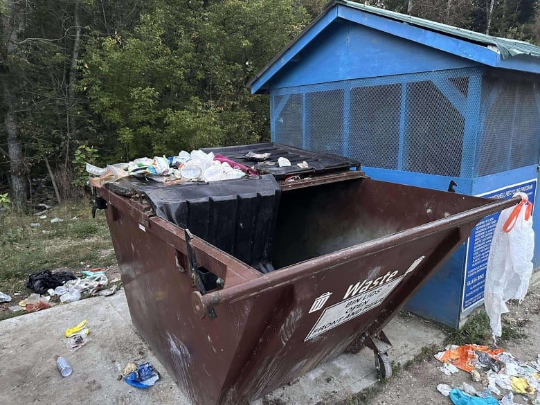 A brown metal garbage bin is overflowing with trash. The lid of the bin is open and damaged. 