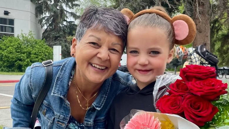 Jill poses with her grandaughter, who wears a headband with bear ears, and holds a bouquet of red roses.