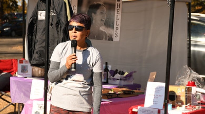 Jillian Young wears sun glasses and running gear as she holds a microphone. A tent with a photo posted of her daughter can be seen in the background