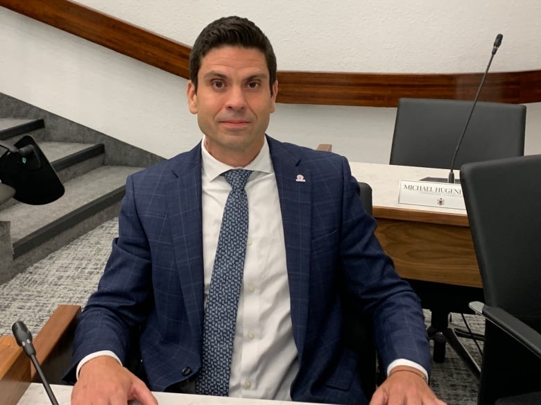 Man wearing a navy blue suit sitting at a table looking at camera with neutral expression. 