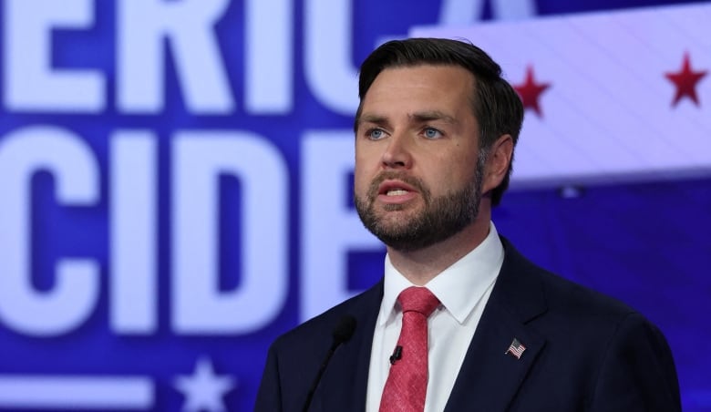 A man in a dark suit with a red tie speaks into a microphone at a podium.