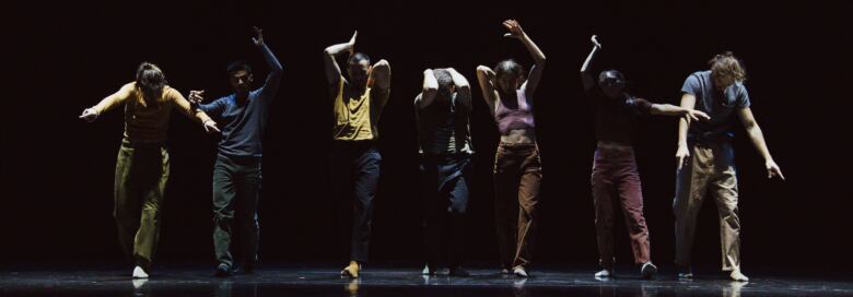 Seven dancers in various poses in a line on stage with a black background.