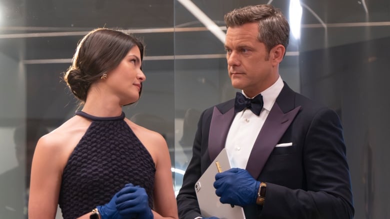 A woman in a black evening gown and blue rubber gloves looks at a man in a black suit and blue rubber gloves.