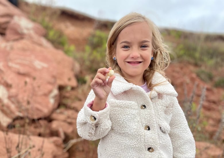 Small blond 6-year-old girl wearing a fuzzy ivory jacket smiles while holding a white beach rock 