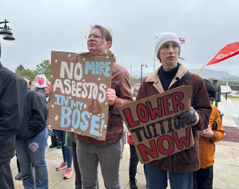 A man and woman hold posters at a protest. The man's sign reads 'No more abestos in my Bose', while the woman's poster reads 'Lower Tuition Now'.