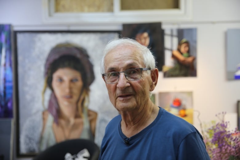 A man with white hair, glasses and a blue T-shirt stands in a home. Paintings are on the walls behind him.