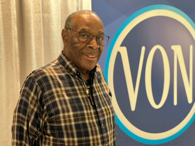 An older Black man smiles at the camera in front of a blue poster that reads 