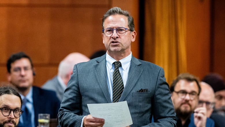 A man in a grey suit holds a piece of paper as he speaks in the House of Commons.