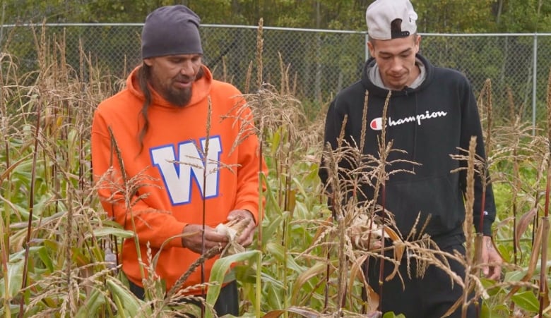 One man wearing a toque and an orange hoodie with a 'W' on it stands screen left to another man wearing a backwards hat and a black Champion hoodie.