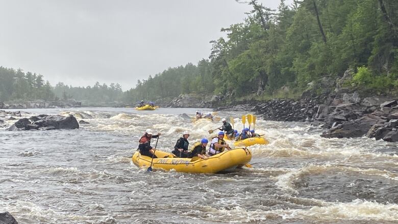 A river is seen on a cloudy, rainy day. There are yellow inflatable rafts in the water fighting the waves. Each raft has four or five people on it, all holding paddles.