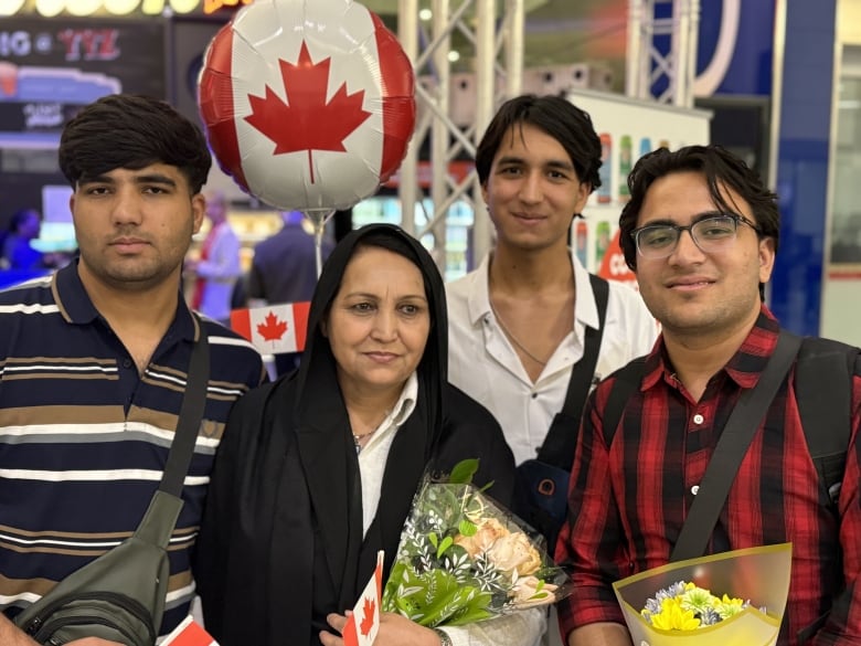 A woman and three young men smile in an airport. They are holding flowers and red-and-white balloons that mirror the Canadian flag.