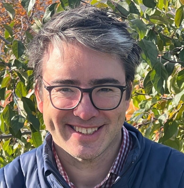 A man with glasses on smiles in front of foliage. 