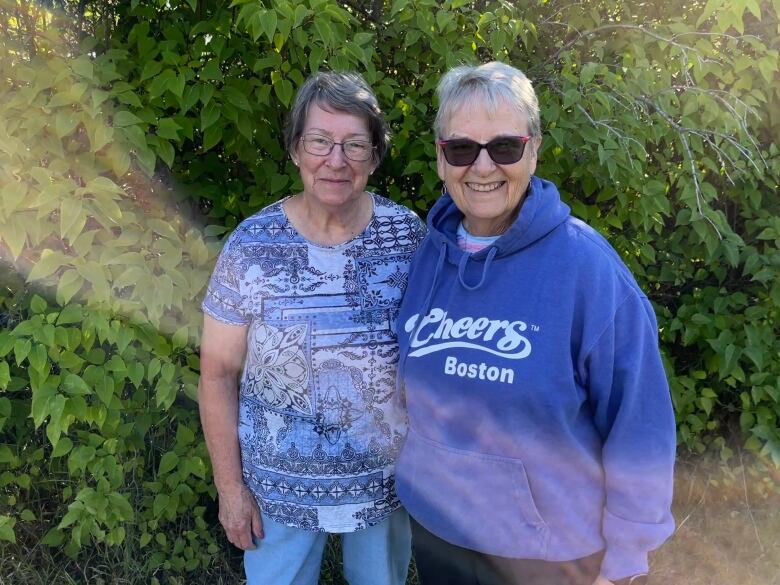 A woman in a blue patterned shirt and he sister in a blue hooded sweatshirt stand in front of greenery on a sunny day.