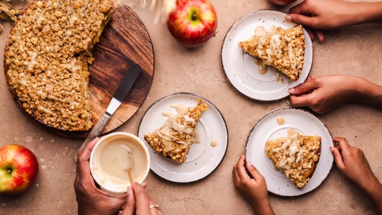 Overhead shot of an apple cake on a wooden cutting board. 3 small plates with slices of cakes are held by 3 sets of hands. 