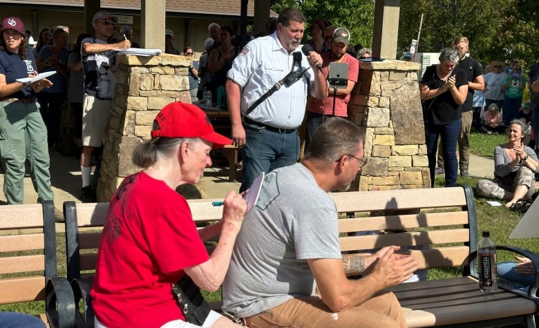 A couple sit on a bench outside. The woman is using the man's back as a desk so she can write on a piece of paper. Beyond them, a man speaks into a microphone. 