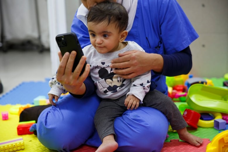 A baby sits on a nurse's laps and looks at a cellphone 