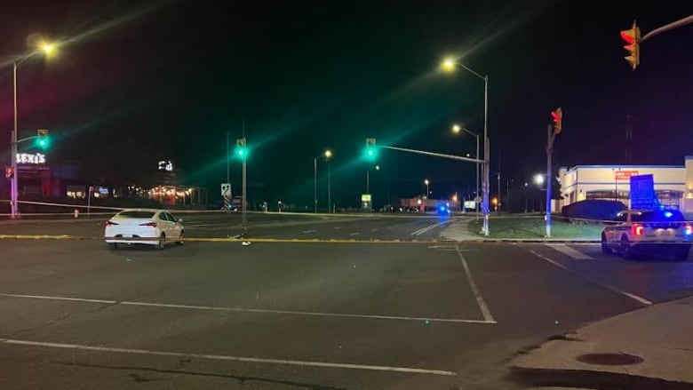 A parked white car near a bicycle on the ground in a city intersection at night. The intersection has yellow tape around it and a police car nearby.