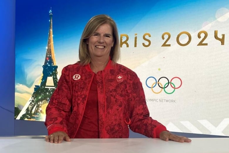 A woman dressed in red stands at a desk and smiles with the Paris 2024 Olympics logo behind her.