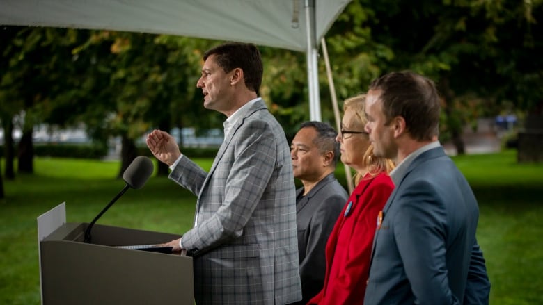 A man in a suit stands at a podium, flanked by two men and one woman in a red suit. 