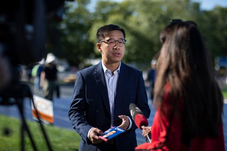 A man in a suite holding an election pamphlet speaks with a reporter.