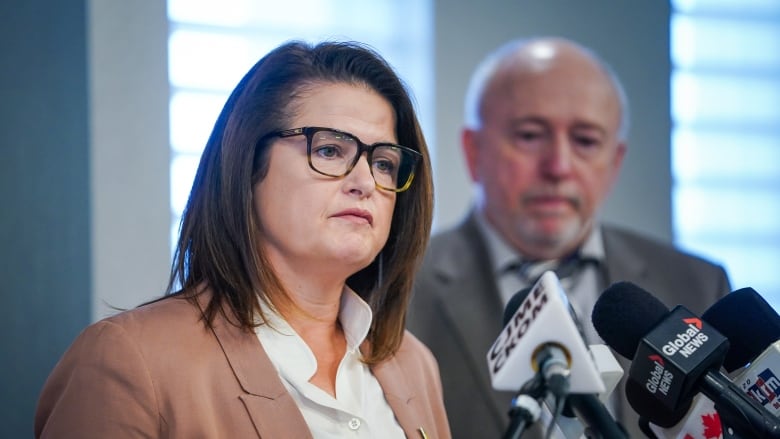 A woman in glasses speaks at a podium. A man in a suit is in soft focus in the background.