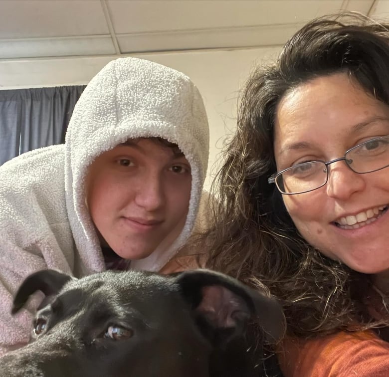 A woman and a young man take a smiling selfie with a black dog.