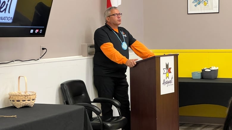 Man dressed in orange shirt with shorter sleeve black shirt over it stands in front of a podium.