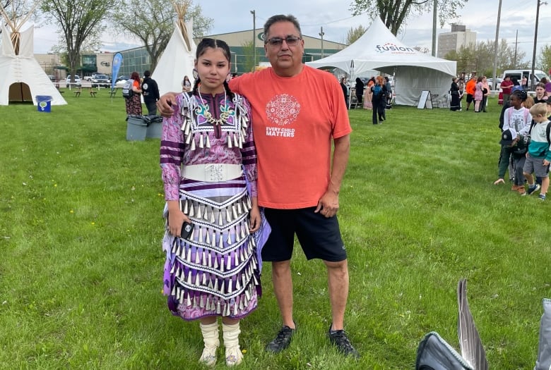 A man in an orange shirt stands with his arm around a young woman in a jingle dress, as they stand in a field in front of tents.